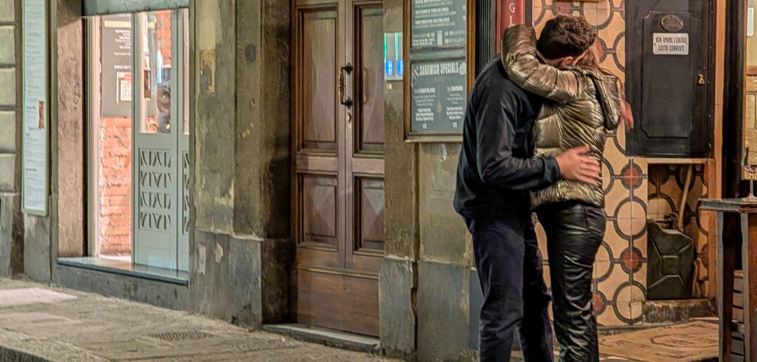 A young couple embraces and kisses intensely on a narrow, dimly lit sidewalk outside the entrance of a restaurant at night in Florence, Tuscany, Italy, on November 22, 2025. The man wears dark clothing while the woman wears a shiny gold puffer jacket and black leather pants. They stand close together, focused only on each other against the backdrop of a wooden doorway and illuminated storefront signage
