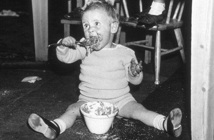 A young child sitting under a table eating Christmas pudding