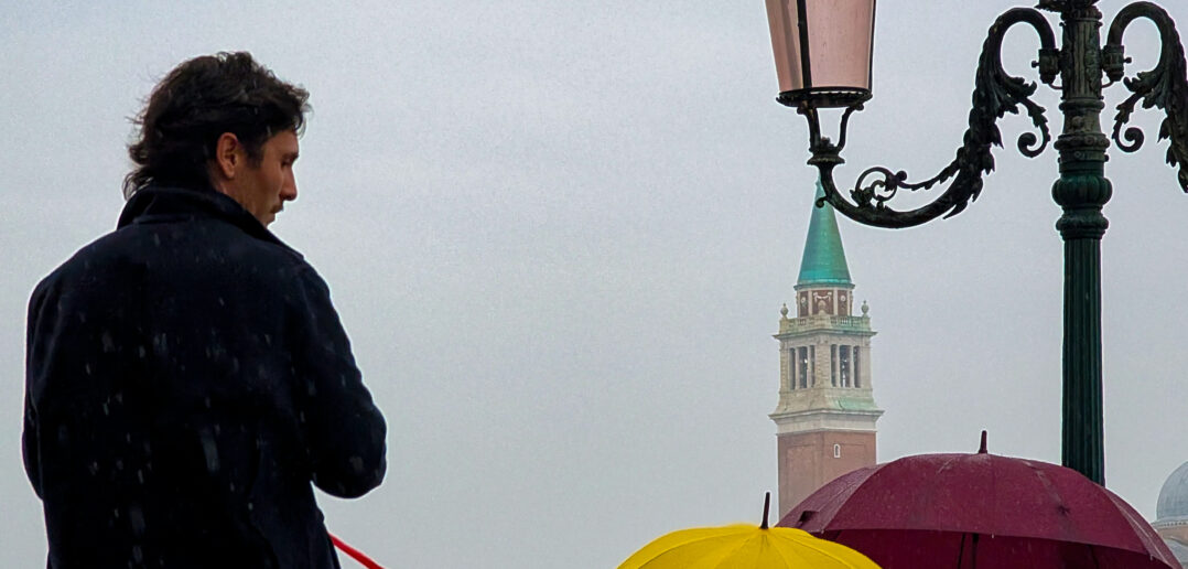A man stands alone near an ornate antique lamppost, looking toward the distant San Giorgio Maggiore bell tower and dome, while other pedestrians shelter under brightly colored umbrellas on an overcast, drizzly day in Venice