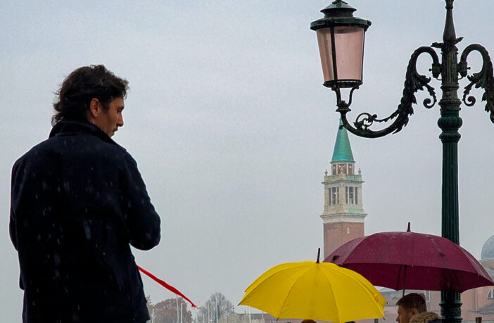 A man stands alone near an ornate antique lamppost, looking toward the distant San Giorgio Maggiore bell tower and dome, while other pedestrians shelter under brightly colored umbrellas on an overcast, drizzly day in Venice